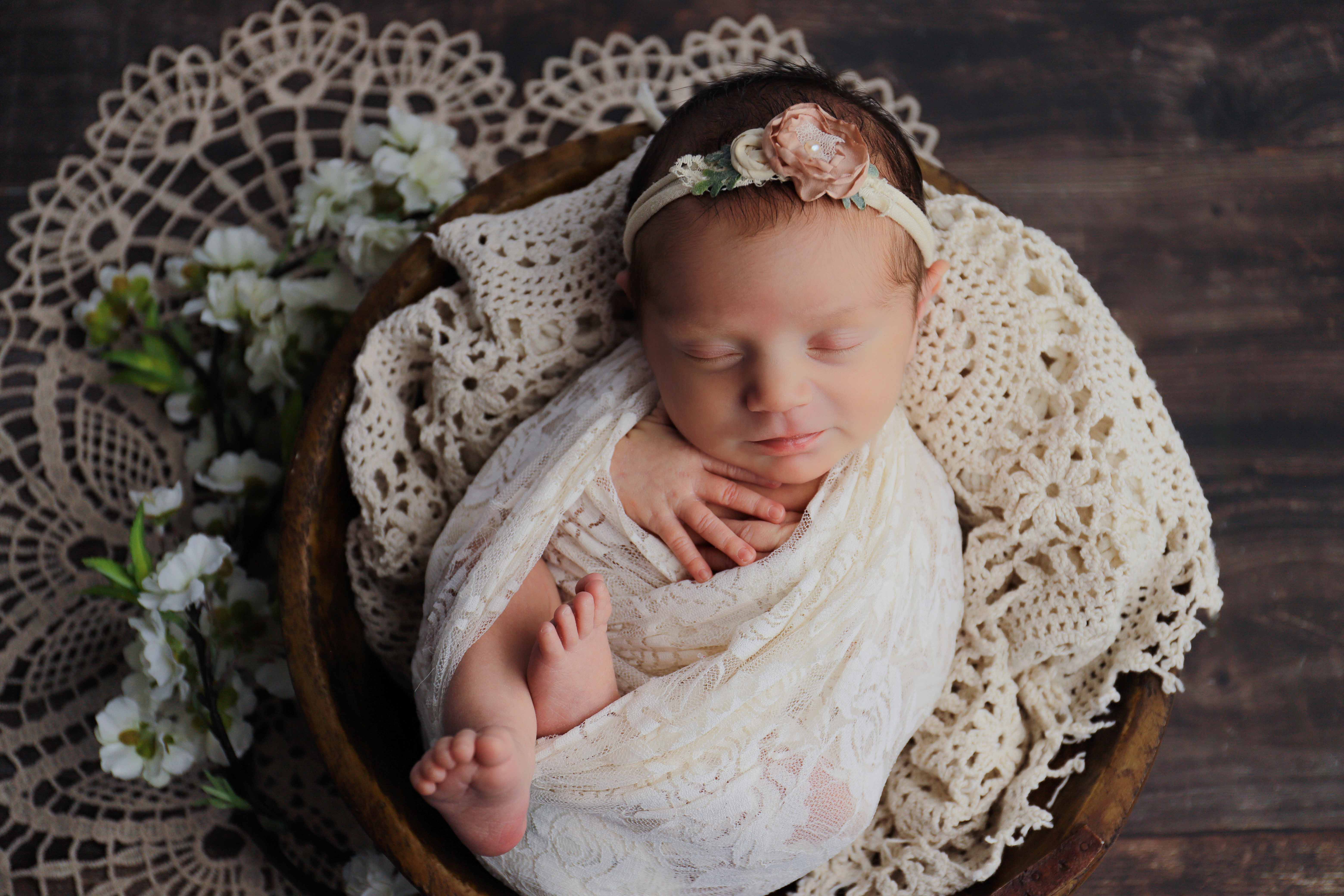 A newborn lying on a textured natural blanket, eyes closed, swaddled in cream.